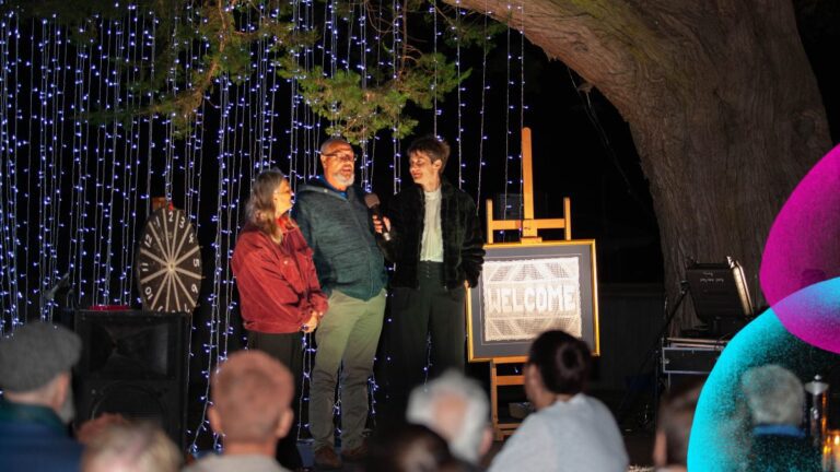 Three people share a microphone on a magical outdoor stage at night, framed by cascading fairy lights and the magnificent canopy of a grand old tree — nature doing what no set designer could. A "Welcome" sign on an easel greets the audience seated in the foreground. Ten Days on the Island festival branding visible on the right.