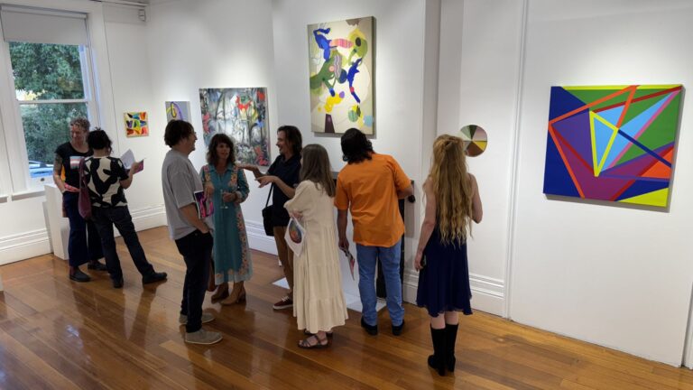Visitors deep in conversation at Colour Rush during Ten Days on the Island 2025, surrounded by paintings so bold they're doing most of the talking. Polished timber floors, white gallery walls, and nobody looking at their phone. Photo: Vernon Guest.