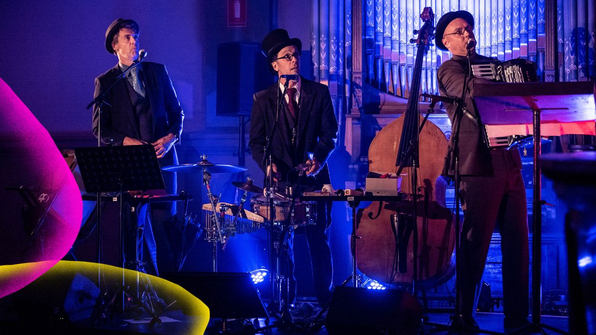 Three sharply dressed musicians perform in a grand heritage theatre bathed in dramatic blue and purple light. All three wear dark suits and bowler or flat-cap hats — a look they've clearly committed to fully. A double bass, accordion and drums fill the stage, with ornate architectural details rising behind them. Ten Days on the Island festival branding visible on the left.