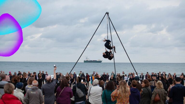 Two aerialists perform an acrobatic duet suspended from a tall tripod rig on a Tasmanian beach, as a crowd of onlookers watches, photographs, and collectively forgets they had morning errands to run. A container ship passes calmly on the horizon, apparently unmoved. Ten Days on the Island festival branding appears in the upper left corner.