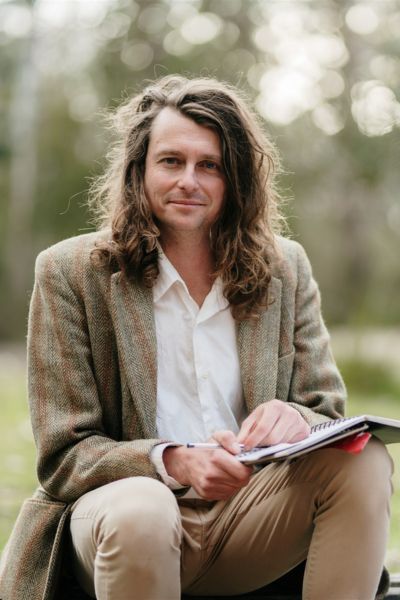 Portrait of writer Bert Spinks, seated outdoors in a tweed jacket, notebook and pen in hand, long curly hair framing a calm, direct gaze — a softly blurred bush backdrop suggesting he does his best thinking outside.
