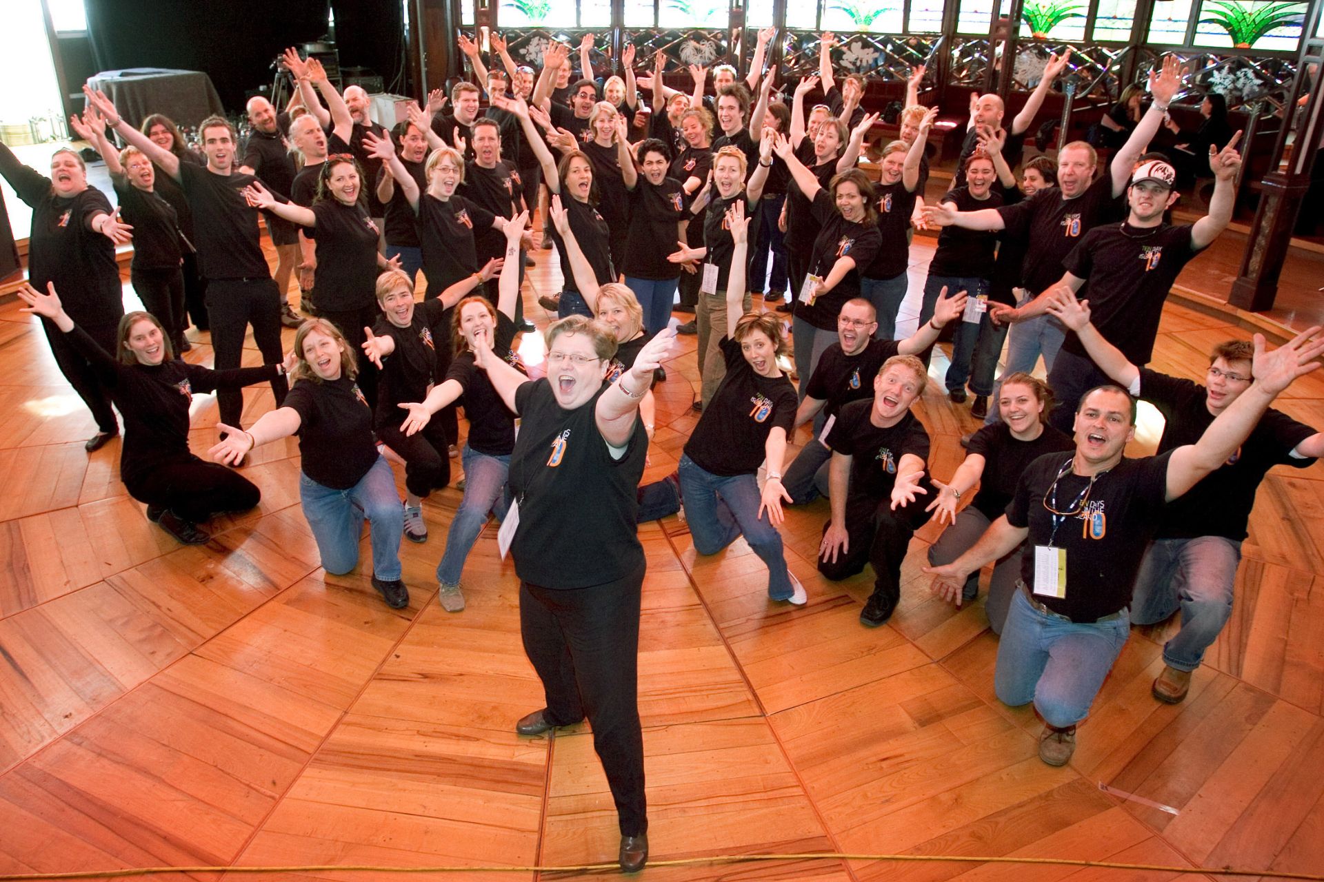 The Ten Days on the Island festival team in matching black t-shirts, arms raised and beaming, photographed from above on a polished timber dancefloor inside a grand ornate ballroom. Robyn Archer leads the charge front and centre with arms flung wide, and absolutely nobody is playing it cool. Circa 2005.