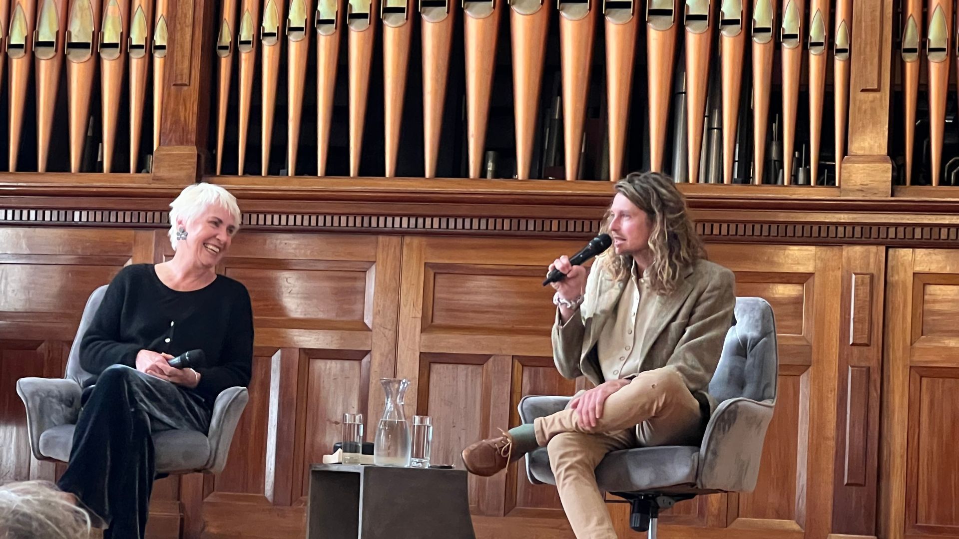 Writers Ailsa Piper and Bert Spinks in conversation on stage, seated in grey velvet chairs before a magnificent wall of timber organ pipes. Ailsa laughs as Bert leans forward mid-thought, microphone in hand — the kind of exchange where the audience forgot to check the time.