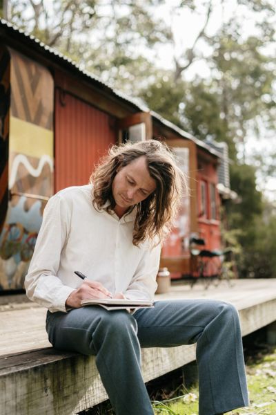 Bert Spinks sits on a weathered timber deck outside a colourful carriage, writing in a notebook, looking exactly like someone who lives in the bush without electricity and is absolutely fine with that.