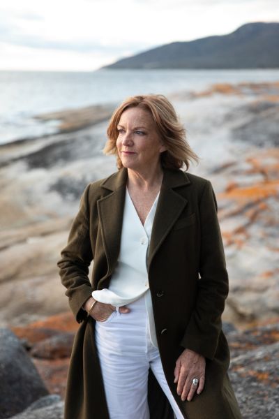 Hilary Burden stands on a rocky Tasmanian coastline, wearing a brown coat over white, her hair catching the breeze. She is looking out to sea with the quiet confidence of someone who has found exactly the right place to think. Mountains and dramatic orange-lichened rocks frame the background.