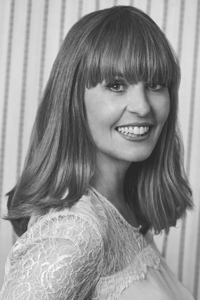 Kirsten Galliott smiling broadly in a lace top, fringe perfectly placed, bringing excellent energy to a striped background.