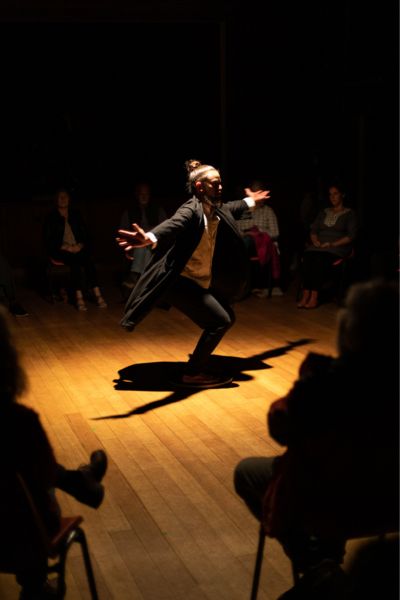 A solo dancer commands a warm spotlight on a timber floor, arms outstretched and knees bent in a moment of pure intention — their shadow giving an equally committed performance just below. A small intimate audience sits in a circle around them, close enough that nobody can check their phone without being noticed. Ten Days on the Island festival.