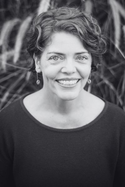 Vanessa Pigrum smiling outdoors with curly hair and drop earrings, tall grasses behind her suggesting she is equally at home in a boardroom or a paddock.