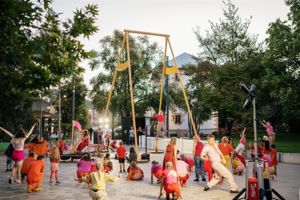 Tasdance performers in a riot of orange, red, pink and yellow costumes take over Civic Square in Launceston for BEACON at the 2025 Ten Days on the Island festival. A towering yellow swing-set installation, SWING, presides over the scene like the world's most artistic playground. A performer stands boldly on the swing seat itself, arms wide, while dancers of all ages crouch, lunge and reach skyward across the paving, some with the grace of professionals, others with the joyful commitment of people who just got swept up in it. A pink harp and musicians hold court to the left, a shipping container with SWING signage anchors the background, and heritage buildings and leafy trees frame it all under a soft Launceston dusk.