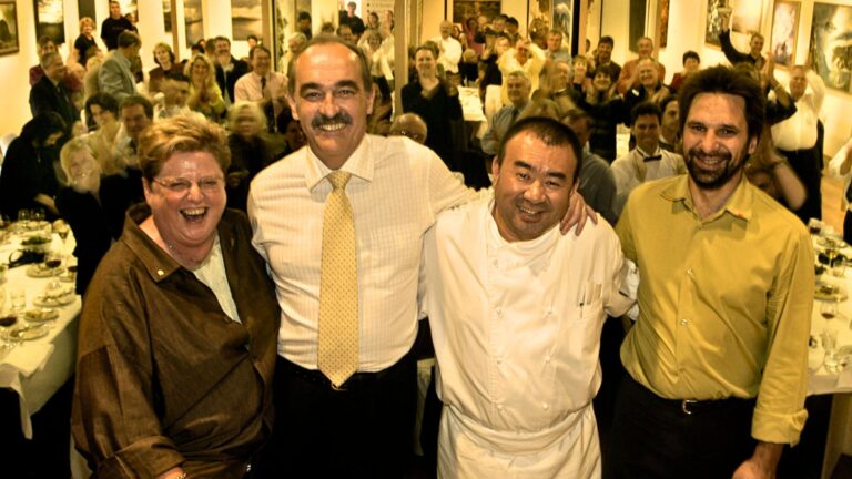 Robyn Archer, Jim Bacon, Tetsuya Wakuda and Mark Bishop stand shoulder to shoulder at the front of a long table dinner during the 2003 festival, with a large seated audience visible behind them in a gallery-style dining space.