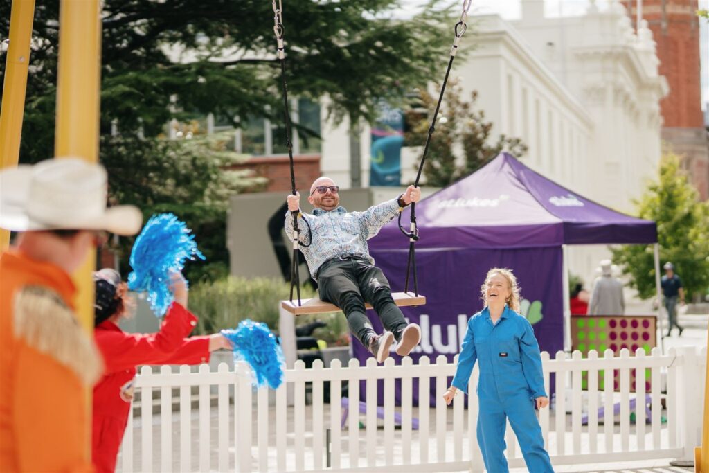 A man is on a giant swing with people standing around in jumpsuits and waving pompoms.