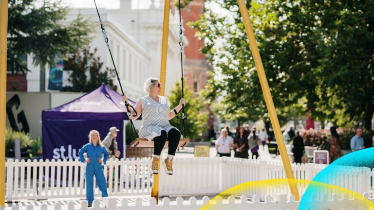 Marnie on SWING with a young woman in a blue jumpsuit standing behind ready to give her a mighty push. Fesitval 2025.