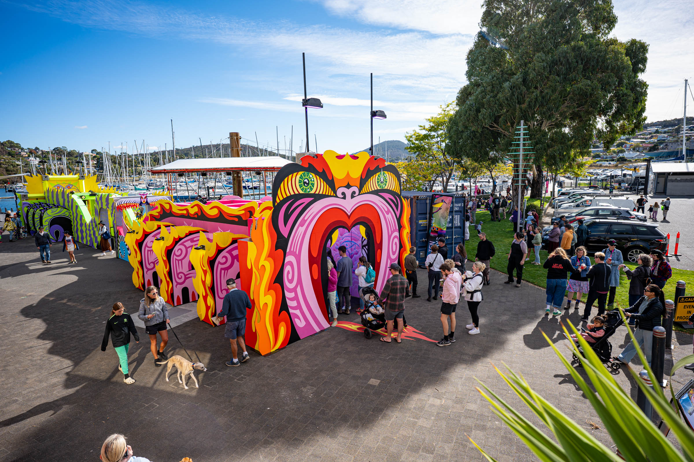 A towering carnival archway painted in pink, yellow and orange swirls inspired by Maori design, set up dockside at a marina. Families and festival-goers wander through and around it in bright sunshine. Ten Days on the Island.